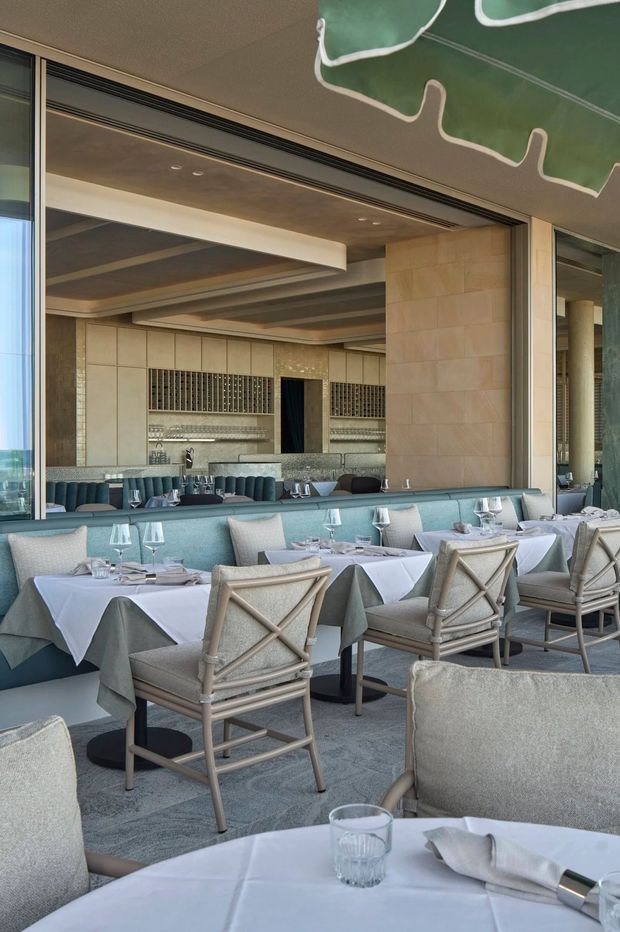 Upscale restaurant dining room with neatly set tables, white tablecloths, upholstered banquette seating, cross-back chairs, and a long bar or service counter in the background. The interior features pale stone or tiled wall surfaces, warm wood-toned ceiling beams, extensive glazing, and a muted blue-grey and beige palette. A sculptural green ceiling installation adds a decorative accent.