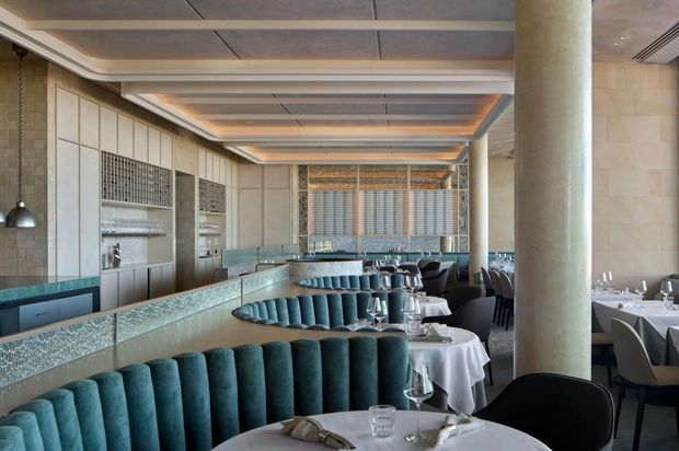 A sophisticated contemporary restaurant dining room with rows of round tables dressed in white linens, teal upholstered banquette seating, dark dining chairs, and a long reflective bar or service counter. The space features large cylindrical stone-toned columns, pale stone wall cladding, decorative grilles, layered recessed ceiling panels with warm concealed lighting, pendant lights, and expansive glazing or mirrored surfaces. The atmosphere is refined and luxurious with a cool teal and neutral stone palette.