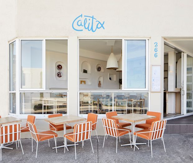 Exterior street-front view of a small contemporary café with large glazed windows and open folding doors. The white façade has blue signage and numbering, while the outdoor terrace is furnished with bright orange-and-white metal chairs and light stone-topped tables. The café interior is partially visible through the glazing, including a counter, shelving, and pendant lighting.