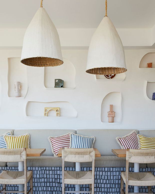Bright, refined living or lounge area with a pale built-in sculptural wall featuring recessed arched niches and display objects. Two oversized white pendant lights with woven undersides hang above a long light-grey upholstered banquette. Several woven dining or counter chairs and striped cushions add a relaxed coastal character, while the lower wall or joinery is finished in a blue-and-white geometric patterned surface.
