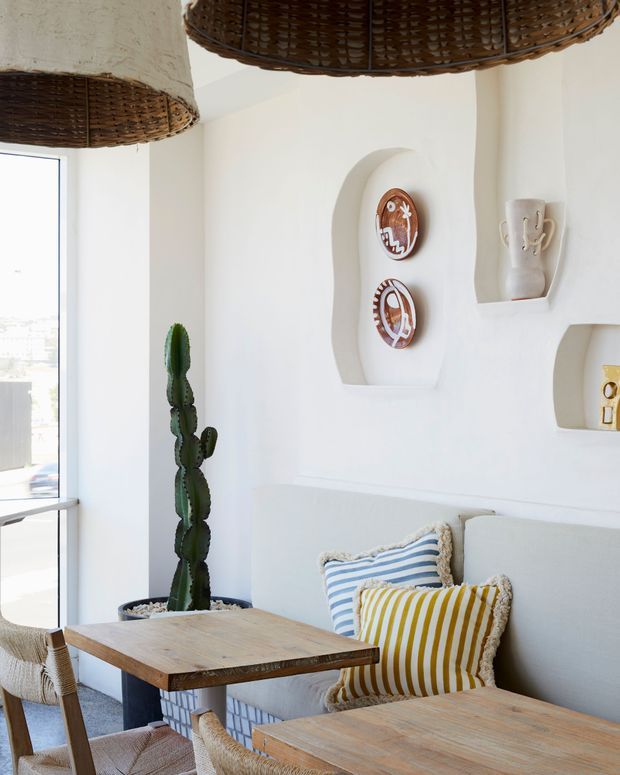 Bright coastal-inspired dining and living area with white sculpted walls, built-in display niches, woven pendant lights, a tall cactus, and a pale upholstered banquette. Rustic timber dining tables and woven chairs are paired with blue-and-white and mustard striped cushions, creating a relaxed, natural interior.