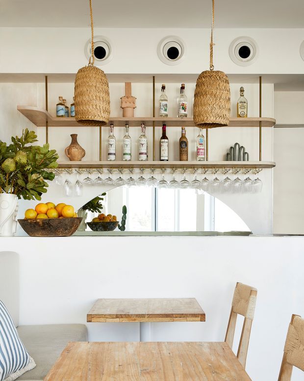 Bright, relaxed dining and home bar area with a white built-in counter, open timber shelving displaying liquor bottles and glassware, woven pendant lights, and a curved mirrored or glazed opening. A timber dining table and chairs occupy the foreground, with natural greenery and fruit adding warmth.
