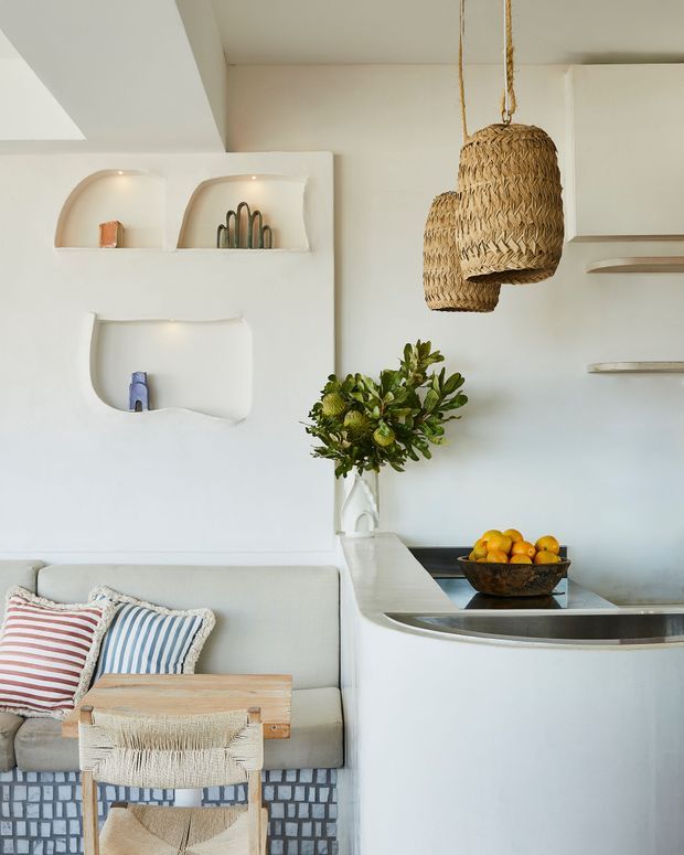 Bright, compact open-plan interior combining a small kitchen or kitchenette with an adjoining living area. The space features sculpted white built-in wall niches, a curved white peninsula with integrated sink, pale cabinetry and surfaces, woven pendant lights, a leafy floral arrangement, and a light sofa with striped cushions. Natural woven textures and soft neutral finishes create a relaxed coastal, organic feel.