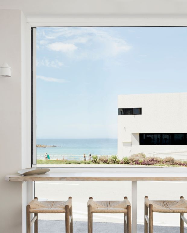 Bright coastal dining or kitchen bar area viewed from indoors through a large picture window. A pale timber counter runs across the foreground with woven timber stools, while the window frames a sandy beachfront, ocean, and a minimalist white flat-roof building. The interior is light, airy, and simply detailed.