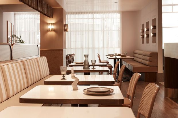 A warm, refined restaurant dining room with rows of cream-and-tan tables, upholstered banquette seating, woven dining chairs, soft sheer curtains, fluted wall details, and sculptural wall lighting. The space has abundant filtered daylight and polished wood flooring, creating an elegant mid-century-inspired atmosphere.