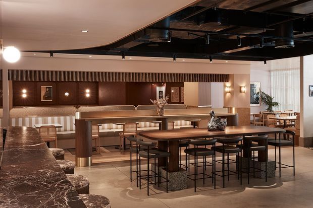 A sophisticated contemporary hospitality-style dining and lounge interior with a long communal timber table, high stools, booth seating, marble-topped counter, and warm brown wall panelling. The space features exposed black ceiling services, track lighting, sculptural decor, and large sheer-curtained windows.