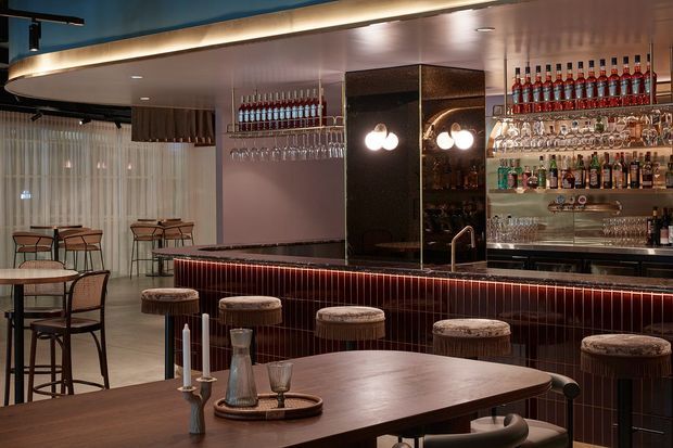 A stylish bar and dining area with a long dark tiled counter, upholstered stools, mirrored shelving, and an extensive display of bottles and glassware. Bistro-style tables and chairs sit beside sheer curtains, while warm integrated lighting and reflective surfaces create an intimate hospitality atmosphere.