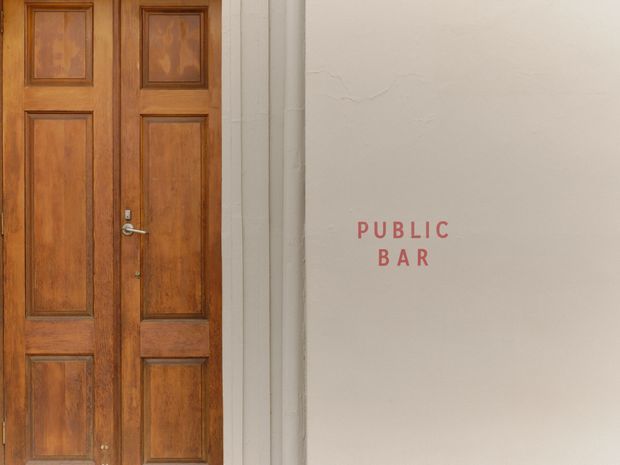 A close-up architectural view of a traditional dark timber double door with raised recessed panels and metal hardware, set beside a pale textured plaster wall. The wall carries simple red lettering reading “PUBLIC BAR,” indicating an entrance to a hospitality venue. The composition is minimal and softly lit, with the door providing the main visual focus.