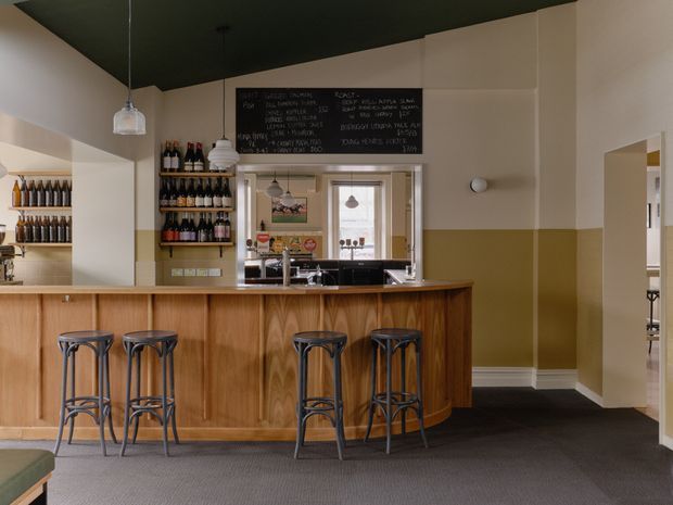 A warmly styled commercial bar or café interior with a curved timber-fronted counter, dark stone or laminate benchtop, and four metal bar stools. Built-in bottle shelving and a chalkboard menu are positioned behind the counter, with a service opening leading to a back preparation area. The room has two-tone cream and mustard walls, a dark ceiling, pendant lighting, and dark carpet.
