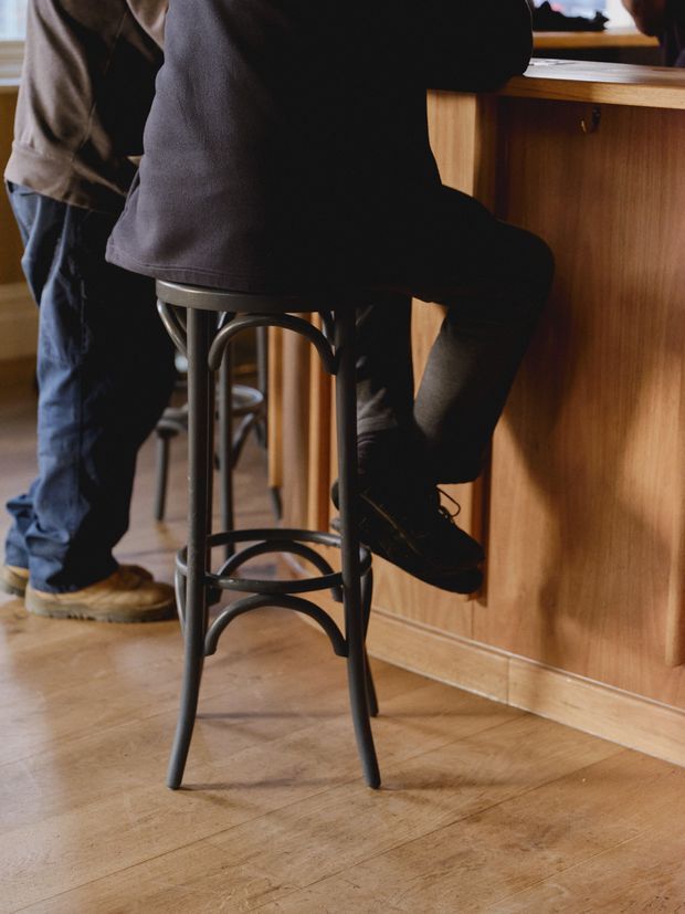 Close-up interior view of a bar or café counter, showing a person seated on a tall black metal bar stool and another person standing nearby. The space features warm timber panelled cabinetry or counter fronts and a light timber floor. The image focuses on the seating and counter area rather than the full room.