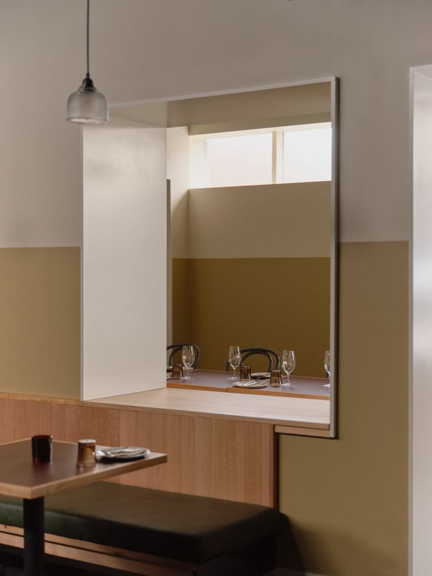 A warmly lit contemporary dining area with a built-in banquette or upholstered bench in the foreground and a timber dining table. A large interior serving opening with a mirrored or reflective surface connects visually to another dining setting, while a simple pendant light hangs above. The room features two-tone beige and ochre painted walls, light timber joinery, and a restrained modern palette.