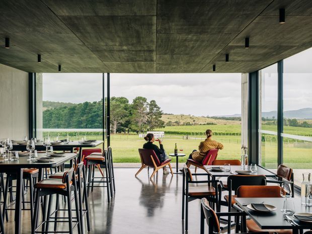 A contemporary dining space with floor-to-ceiling glazing overlooking open green farmland and rolling hills. The room features a dark exposed concrete ceiling with recessed cylindrical lights, polished concrete flooring, slim dark window frames, and numerous black-framed dining tables and chairs with warm timber or leather seating. A small seating area sits near the glazing.
