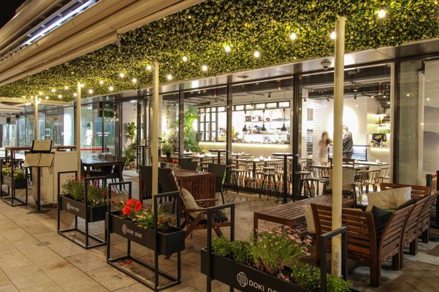 A covered outdoor dining terrace adjoining a modern café or restaurant. The space features timber tables and chairs, black metal planter boxes with greenery and red flowers, polished stone paving, slim structural columns, glass walls, and a dense green ceiling canopy with warm pendant lights. Indoor dining and bar seating are visible through the glazing.
