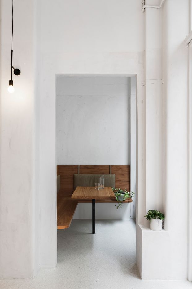 A minimalist interior viewed through a wide doorway, featuring a built-in timber booth with upholstered seating, a small timber table on a black central pedestal, white plaster walls, exposed white-painted pipework, and pale polished flooring. A simple pendant light and potted greenery add restrained accents.