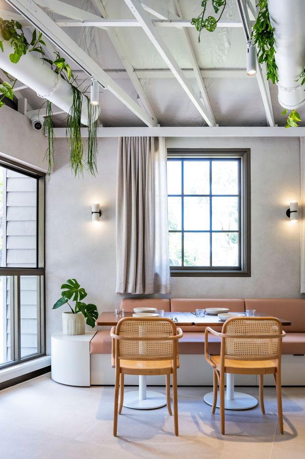 A bright contemporary dining area with a built-in blush-toned banquette, two cane-backed timber dining chairs, and a compact round dining table. The room has pale tiled flooring, textured light walls, large dark-framed grid windows, exposed white-painted ceiling beams and services, wall sconces, soft curtains, and hanging greenery.