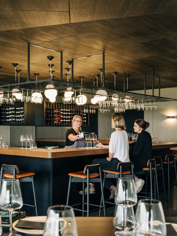 A contemporary wine bar interior viewed across a dining table toward a dark bar counter. The space features extensive suspended glassware racks, illuminated pendant lights, wall-mounted wine displays, black cabinetry and bar stools with warm timber seats. A bartender serves two seated patrons, while wine glasses and table settings occupy the foreground. The ceiling and portions of the walls are clad in dark, warm-toned timber, creating an intimate hospitality atmosphere.
