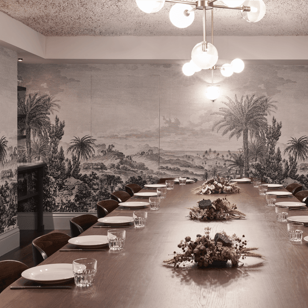 A refined dining room with a long timber dining table set for a large gathering, surrounded by dark curved chairs and white ceramic plates with glassware. A large monochromatic scenic mural depicting palms, vegetation and a distant landscape covers the walls, while sculptural globe pendant lighting hangs above. The room has a warm, elegant, atmospheric character with a textured ceiling and dark timber flooring.