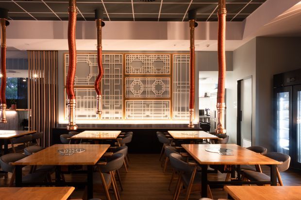 A warmly lit restaurant dining room with multiple timber tables and dark upholstered chairs. A large illuminated geometric decorative screen forms the feature wall, with vertical timber slats to one side. Exposed copper pendant ducts hang from a black grid ceiling, while dark timber flooring and a partially visible bar or service area reinforce the hospitality setting.