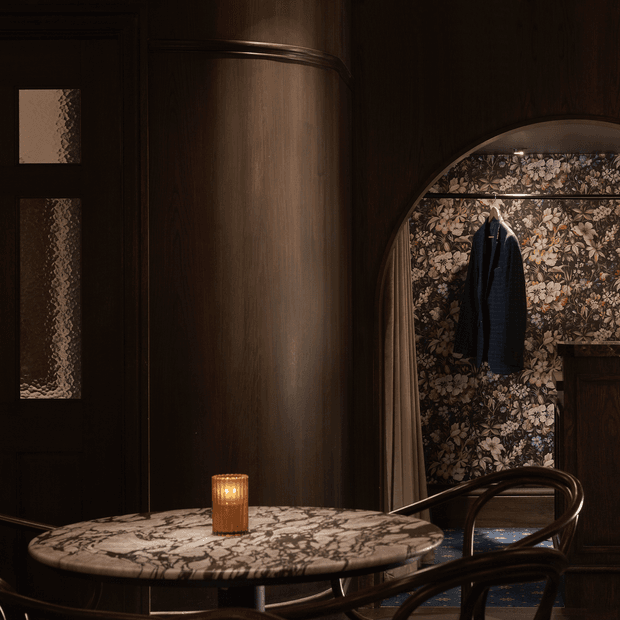 A dark, atmospheric interior vignette featuring a round marble-topped table with a candle, curved dark timber wall panelling, a textured glass door, and an arched alcove lined with floral wallpaper and a hanging coat. Rich timber tones, patterned surfaces, and low warm lighting create a moody, refined hospitality-style setting.