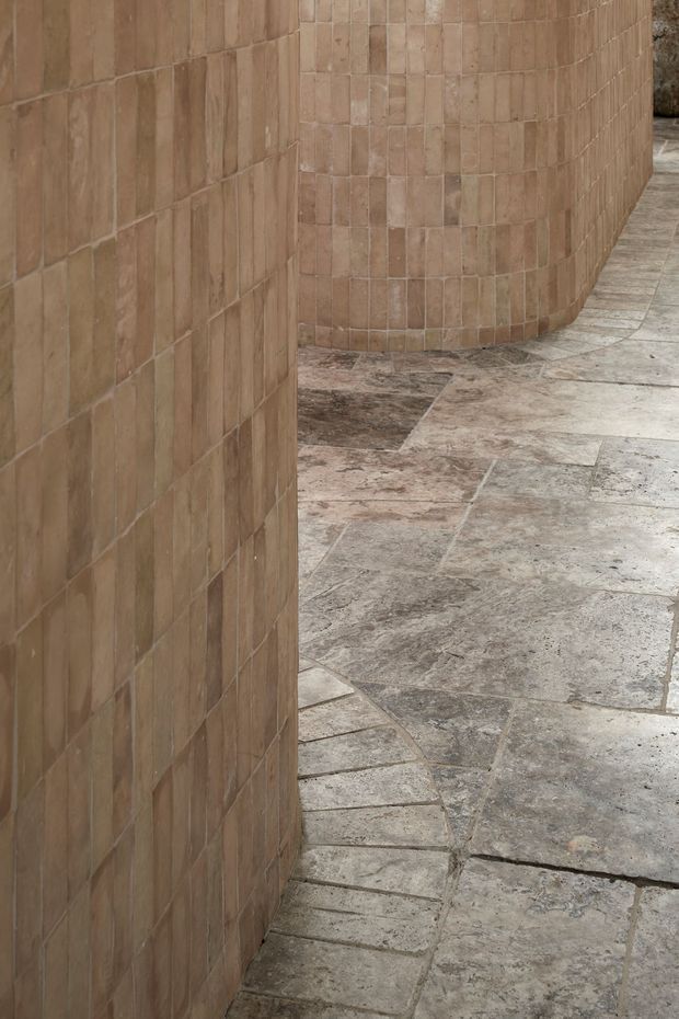 Close-up architectural detail of a curved wall and adjoining straight wall clad in small rectangular terracotta-toned tiles, with a worn irregular stone-paved floor following the curved passage. The image focuses on tactile masonry and paving materials, with no furniture or people visible.