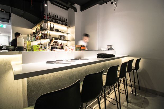 A compact contemporary commercial bar or café interior with a long pale counter, black bar stools, illuminated textured front panel, and a back bar displaying bottles and glassware on open shelves. The space has white walls, dark exposed ceiling services, tiled flooring, and focused warm lighting.
