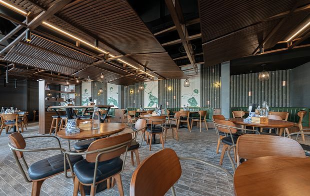 A spacious contemporary commercial dining venue or restaurant with numerous round timber tables, upholstered and timber-framed chairs, a long bar and shelving area, and decorative pendant lighting. The interior features dark ribbed wall panels, extensive timber slatted ceiling treatments, exposed dark structural beams, concrete-look flooring, and patterned mural panels behind the bar. The atmosphere is warm, refined, and slightly moody.