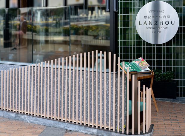 Exterior storefront frontage for a beef noodle bar, featuring a curved low screening wall with closely spaced pale timber slats, dark speckled infill, black-framed reflective glazing, and a dark green tiled facade. A circular sign identifies the venue as Lanzhou Beef Noodle Bar. Brick paving and a small outdoor seating area complete the street-facing composition.