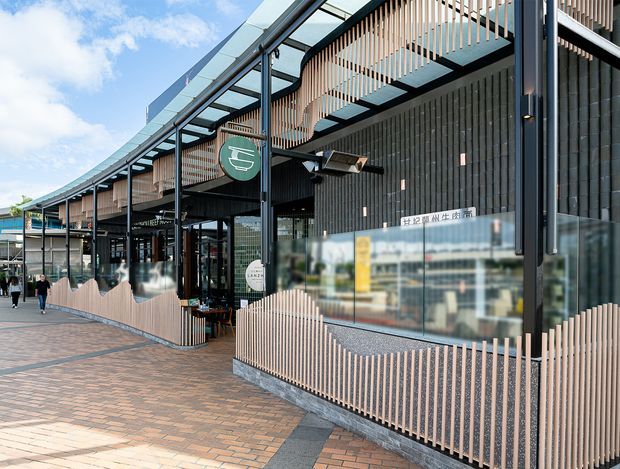 Exterior view of a contemporary commercial building with a long covered pedestrian frontage. The facade combines dark vertical cladding, extensive glazing, black steel framing, and repeating pale timber-look vertical fins used as screens and balustrades. Brick-paved paving extends along the storefront, with signage and entrances visible beneath the canopy.