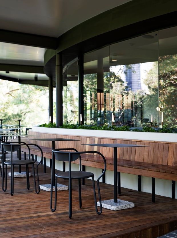 A contemporary café or hospitality dining area with black metal café tables and chairs arranged along a built-in timber bench. The space features a timber deck floor, full-height glazed folding or sliding doors, a long white planter with greenery, dark structural columns and beams, and views into a leafy outdoor setting. The fit-out has a refined indoor-outdoor character with warm timber and dark metal detailing.