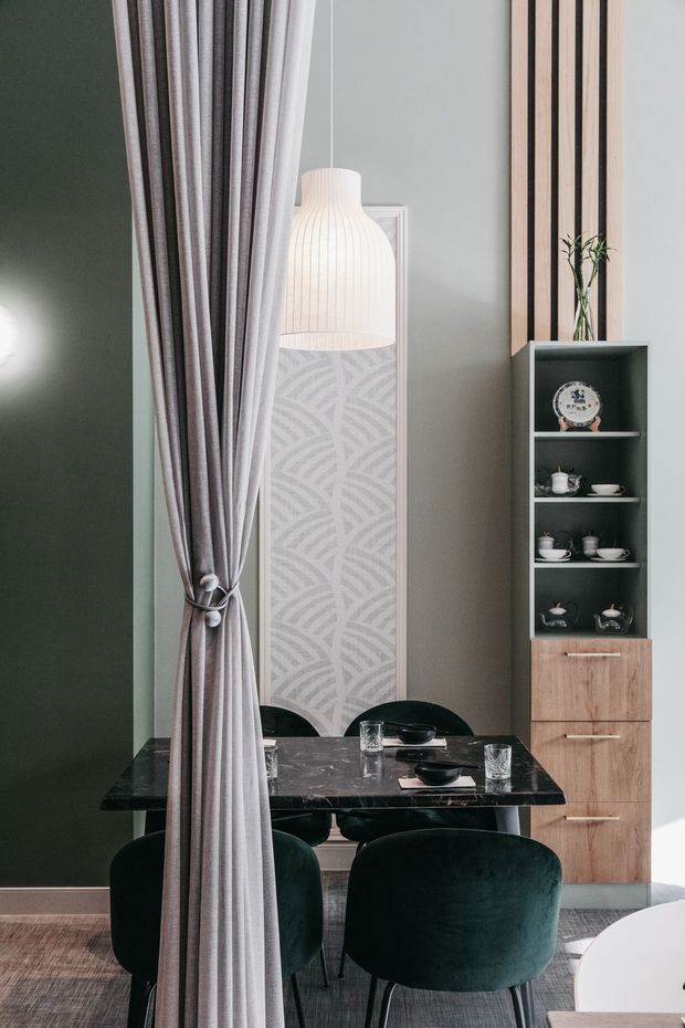 A sophisticated dining area viewed through a partially drawn floor-to-ceiling curtain. The room features a dark marble dining table with dark upholstered chairs, a tall patterned floor lamp, a built-in timber and dark open-shelving unit displaying tea ware, vertical timber wall battens, muted green and grey walls, and soft neutral textiles. The styling is refined and contemporary with a restrained, moody palette.