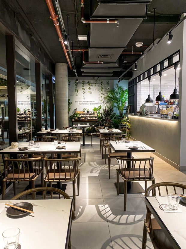 A contemporary restaurant dining interior with rows of neatly set square tables and wooden dining chairs. The space features a long illuminated service counter with pendant lights, black-framed glazing, exposed black ceiling services with copper pipes and ductwork, concrete columns, pale tiled flooring, and a lush planted feature wall at the rear.