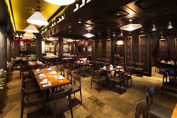 A warmly lit restaurant dining interior with numerous timber tables and upholstered chairs, patterned dark partition screens, decorative wall panelling, and pendant lighting. The space has a rich, atmospheric palette of dark wood, bronze tones, and warm amber illumination.