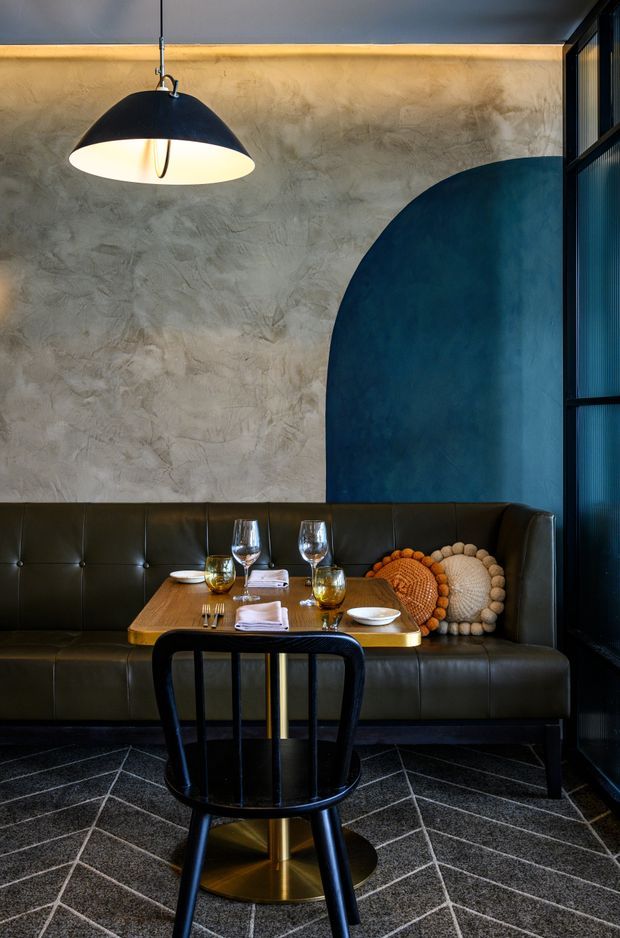 A moody restaurant booth with dark green-black leather banquette seating, a small timber dining table, black spindle-back chair, patterned dark carpet, and warm pendant lighting. The textured plaster wall features a large curved deep-blue painted accent shape, with black-framed ribbed glazing along the right side. Table settings and decorative round cushions create a refined, intimate dining atmosphere.