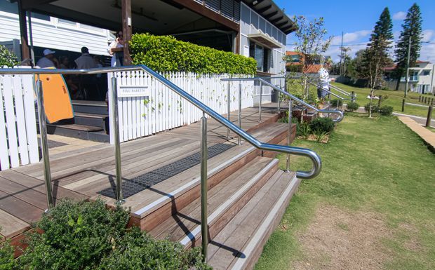 Exterior view of a raised building entrance with broad timber decking and steps, stainless-steel handrails, and an accessible-looking ramp or stair approach. A white vertical-slat facade and covered outdoor seating area sit beside landscaped lawn and a suburban streetscape.