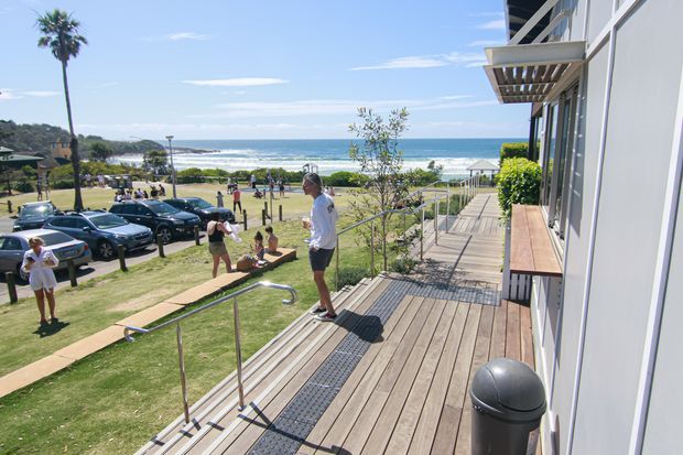 A sunny coastal exterior view from a raised timber deck or access platform beside a modern building. The image looks toward a beach and ocean, with landscaped grass, a pedestrian path, parked cars, visitors, stainless-steel handrails, and an accessible-looking ramp or stepped walkway. The building has light-coloured vertical cladding, large glazing, and a projecting timber sunshade.