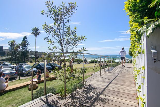 A sunny elevated timber boardwalk or deck with stainless-steel railings, overlooking a landscaped public or residential setting and the ocean beyond. A white-clad building edge with lush greenery frames the right side, while people and parked cars appear in the background.