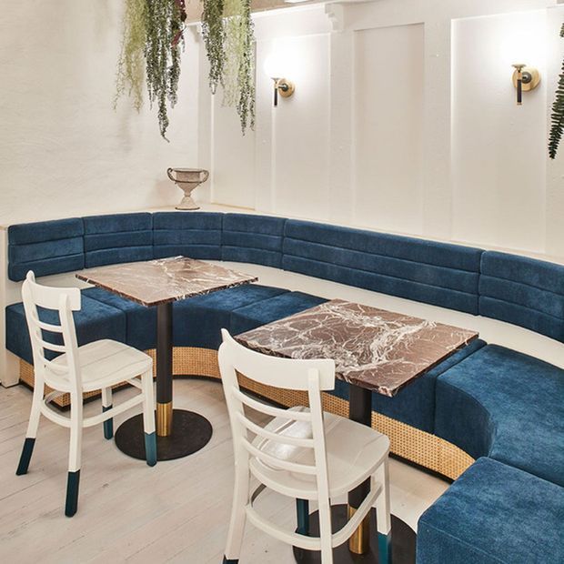 A refined dining or café-style seating area with a curved deep-blue upholstered banquette wrapping around two marble-topped tables. White ladder-back chairs with woven cane details sit on a light floor, while white paneled walls, brass wall sconces, and hanging greenery create a bright, elegant interior.