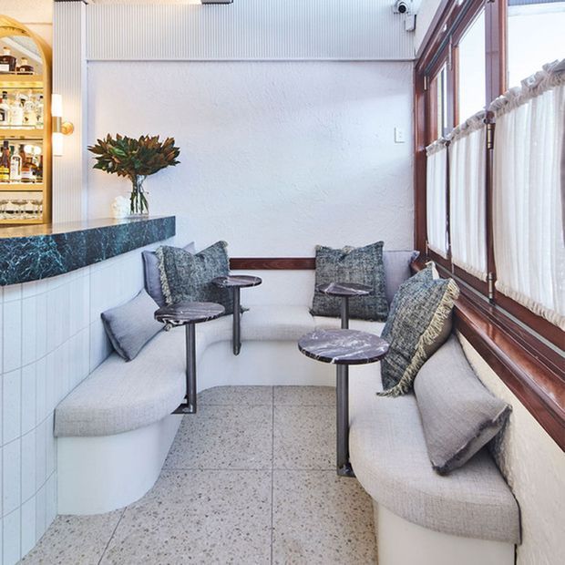 A compact, bright hospitality seating nook beside a bar, featuring curved upholstered banquette seating, small round pedestal tables, textured cushions, a dark blue-green stone bar benchtop, white tiled cabinetry, terrazzo flooring, and large timber-framed windows with sheer curtains. A built-in drinks display is visible at the left.