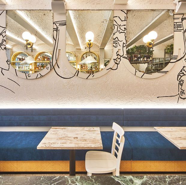 A stylish hospitality interior, likely a restaurant or café, featuring a long blue upholstered banquette, pale wood and black pedestal tables, white chairs, and a textured cream wall with three large rounded mirrors. The mirrors reflect a well-stocked bar, pendant lighting, and decorative greenery. Black line-art detailing frames the mirrors, while a dark veined stone surface appears in the foreground. The overall design is eclectic and refined with Art Deco-inspired curves and graphic accents.