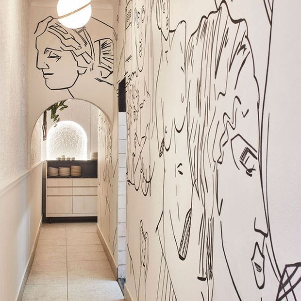 A narrow, softly lit interior hallway with pale tiled flooring and white walls covered in bold black line-art murals. An arched opening leads to a compact timber-toned vanity or storage area, with a round pendant light and decorative mirror adding warmth and character.