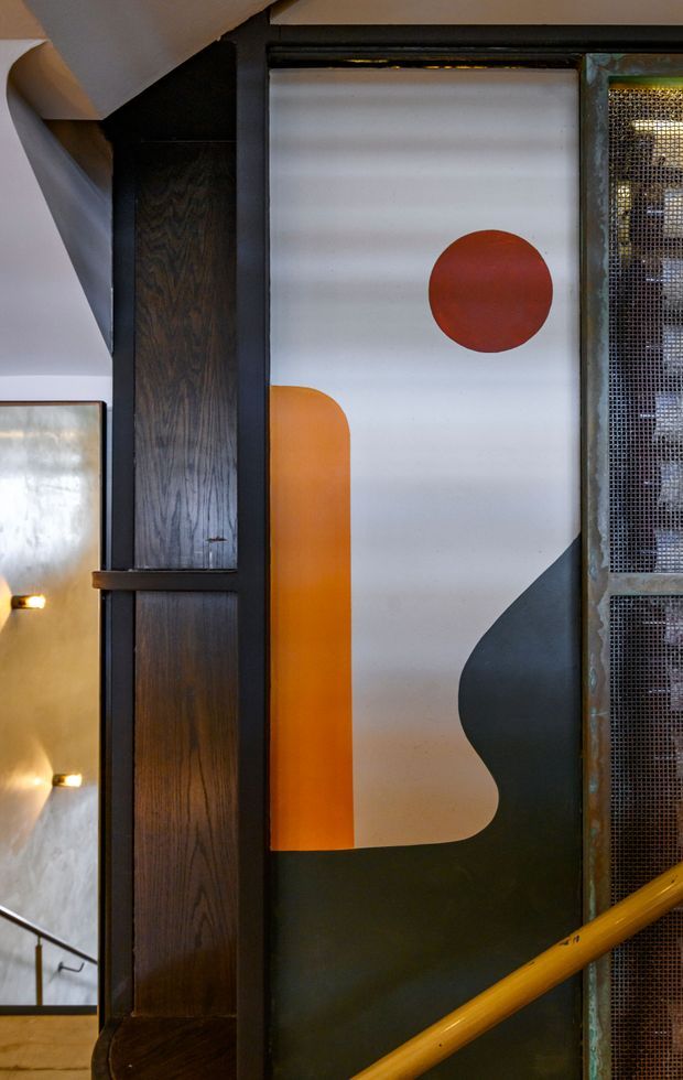 Interior architectural detail showing a stairway or landing with a bold geometric painted wall panel, dark timber vertical joinery, black metal framing, mesh screening, and a mustard-yellow handrail. The composition has an industrial, graphic character with warm low lighting.