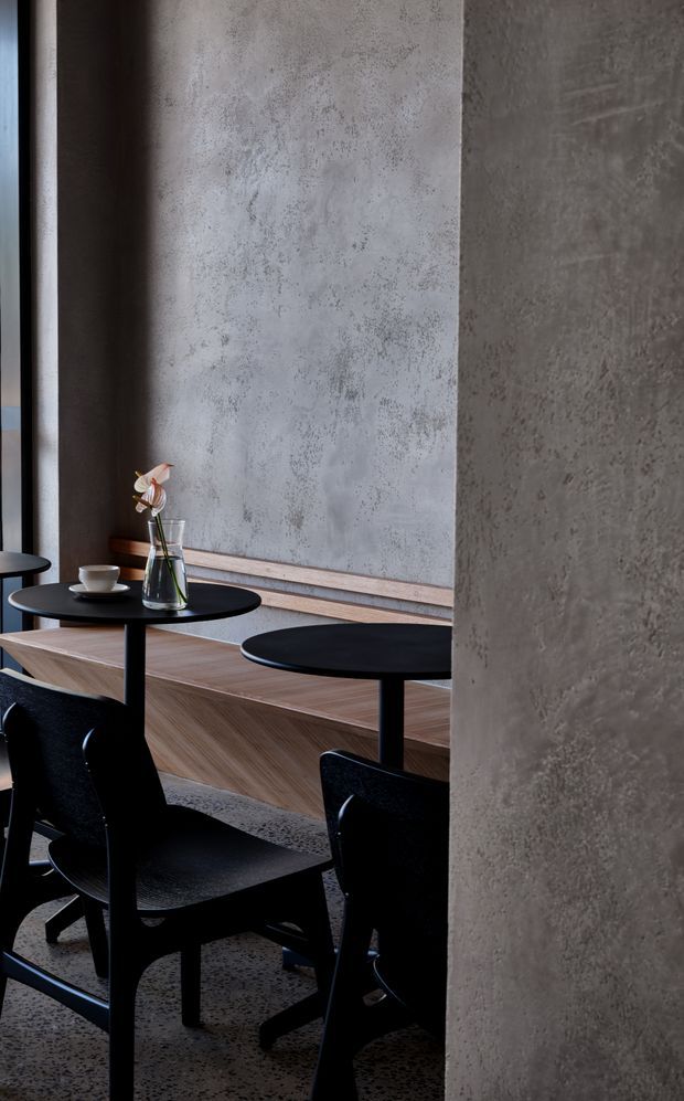 A moody, refined dining or café interior viewed beside a large textured concrete column. The space features black round pedestal tables and sculptural black chairs, a long warm timber banquette or raised joinery element, grey textured plaster/concrete walls, and a small vase with a flower on one table. The composition emphasizes layered vertical surfaces, dark furniture, and a restrained material palette.