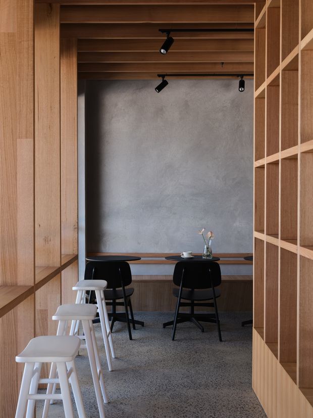A compact, refined café-style seating area framed by extensive warm timber joinery. Built-in timber shelving and wall panels surround a grey plaster/concrete-look feature wall, with a timber slatted ceiling and black track lighting. A narrow counter with black chairs and white stools creates a casual dining or hospitality setting. The floor appears to be grey terrazzo or polished concrete.