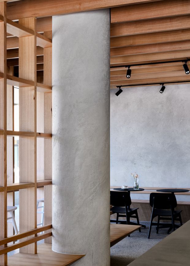 A refined contemporary dining or café-style interior featuring warm timber ceiling slats, open timber shelving, a tall curved textured plaster/concrete-like partition, and a pale textured feature wall. Black track lighting and black timber dining chairs contrast with the warm wood finishes. The space feels calm, minimalist and naturally lit.