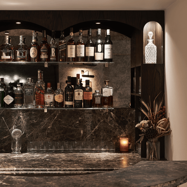 A sophisticated built-in home bar with dark timber arched shelving, illuminated glass shelves, a textured stone or marble backsplash, and a matching veined stone countertop. The shelves display liquor bottles and glassware, while a crystal decanter, candle, and dried floral arrangement add decorative detail. The space has a moody, luxurious atmosphere with warm accent lighting.