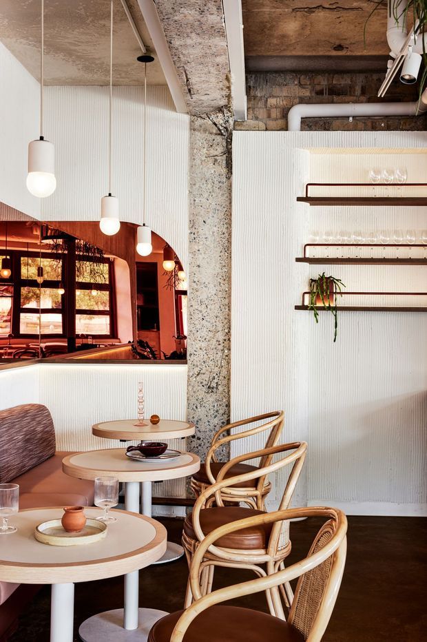 A warmly lit hospitality interior, likely a café or restaurant, with a lounge seating area featuring curved rattan chairs, upholstered banquette seating, round tables, pendant lights, and wall-mounted shelving. The space combines ribbed white wall panels with exposed aged concrete, brick, timber-toned ceilings, visible services, and a richly coloured arched opening leading to a glazed dining area. Plants and glassware add decorative detail.