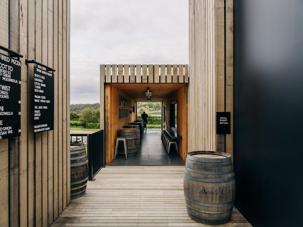 A covered timber-clad passage or entrance to a rustic winery or hospitality venue. Vertical timber battens frame a warm wood-lined tunnel with a timber deck, barrel tables, bench seating, menu boards, and a distant view of landscaped rural surroundings. The space has a restrained contemporary-rustic character with dark accents and practical hospitality fixtures.