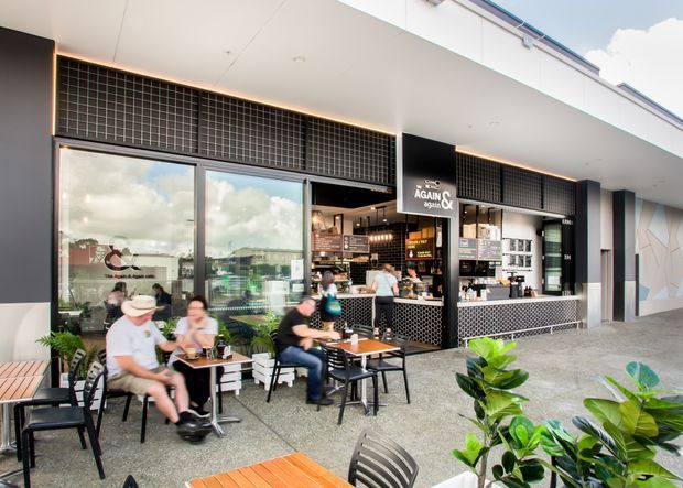 Street-facing café or casual dining venue with a covered outdoor seating area, large glazed shopfront, black tiled façade detailing, patterned counter front, and prominent café signage. Customers sit at timber tables surrounded by potted greenery, while the interior service counter is visible through the open frontage.