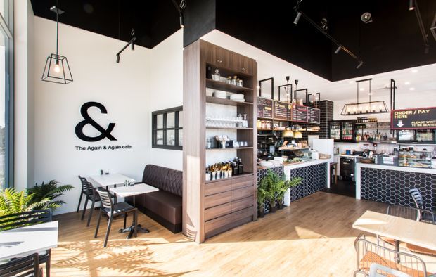 Bright modern café interior with a prominent black ceiling, white walls, warm timber flooring, black-framed windows, pendant and track lighting, banquette seating, café tables, and a timber display shelving unit. The service counter features dark patterned tile, with menu boards and food displays visible in the background.