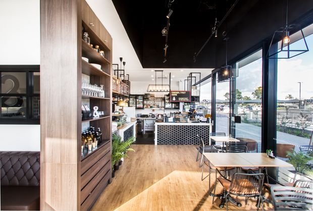 Bright contemporary café interior with a long open service counter, black-and-white patterned counter front, timber flooring, extensive timber shelving and cabinetry, black ceiling with track lighting, and full-height glazed frontage overlooking the street. Dining tables and chairs line the window side, while display shelving and café equipment occupy the service area.